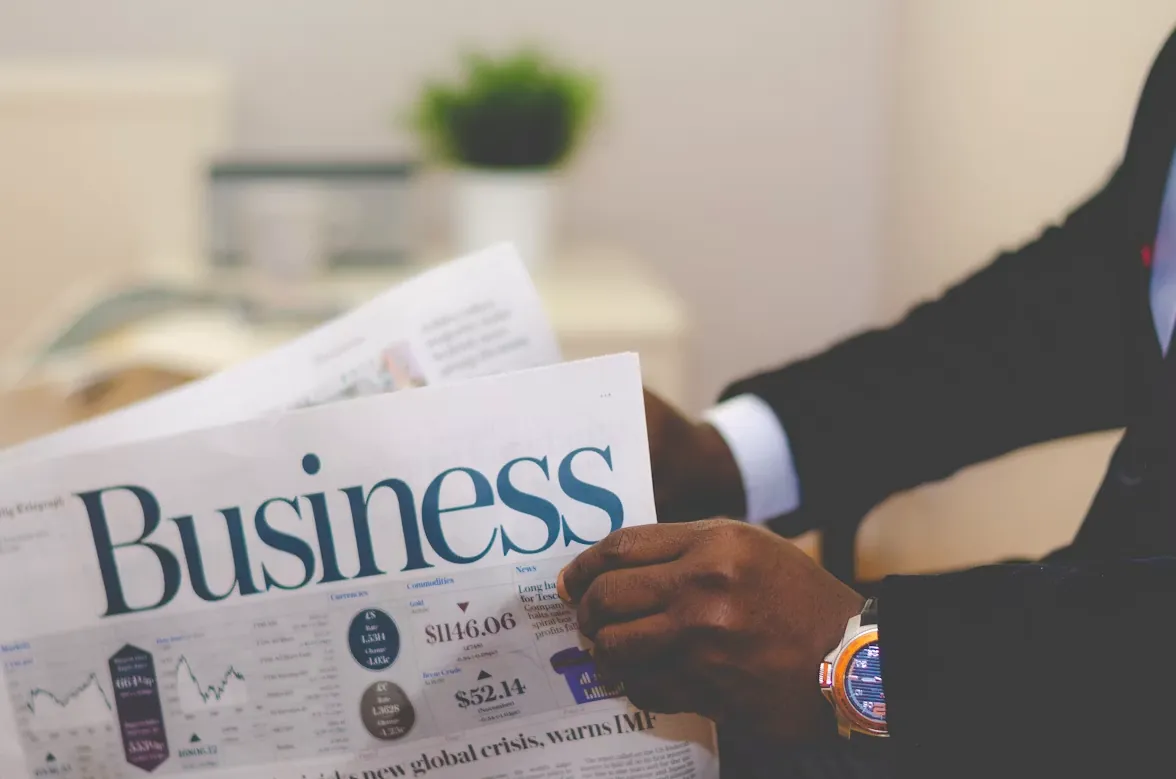 Man in suit reading a newspaper with 'Business' headline, coffee and plant nearby